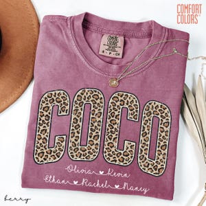 May include: A berry-colored t-shirt with the name "COCO" in large, leopard print letters. Below the name are smaller names: Olivia, Kevin, Ethan, Rachel, and Nancy. A gold necklace and a Comfort Colors tag are visible.
