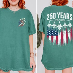May include: A sage green short-sleeve t-shirt featuring a small graphic on the front left chest of a distressed American flag with fighter jets. The back displays the text 250 YEARS 1776-2026 above five fighter jets and a large distressed American flag.
