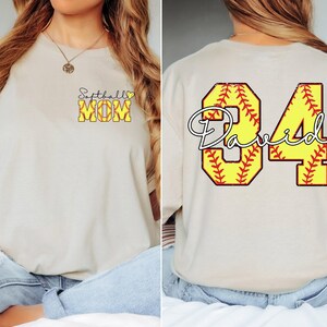 Puede incluir: Camiseta beige con texto "Softball Mom" en la parte delantera y un gr&aacute;fico de softbol. La parte trasera presenta un gr&aacute;fico de softbol amarillo con el n&uacute;mero "84" y el nombre "David".
