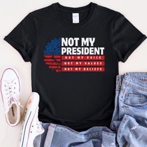 May include: Black t-shirt with a red, white, and blue sunflower graphic and the text "Not My President, Not My Voice, Not My Values, Not My Beliefs".