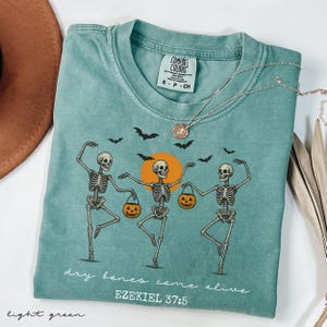 May include: Light green Comfort Colors t-shirt featuring a Halloween design. The graphic shows dancing skeletons holding jack-o'-lanterns, with bats and a sun. The text reads "dry bones come alive" and "EZEKIEL 37:5".