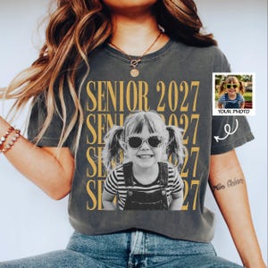 May include: A charcoal gray short-sleeve t-shirt featuring the repeating gold text SENIOR 2027 and a central black and white photograph of a child with pigtails wearing sunglasses and overalls. A small inset box shows the original color photo.