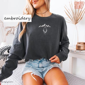 May include: Dark gray long-sleeve sweatshirt with a white embroidered penguin and mountain design. The word "embroidery" is printed on the left side of the sweatshirt. The sweatshirt is made of a soft, comfortable material.