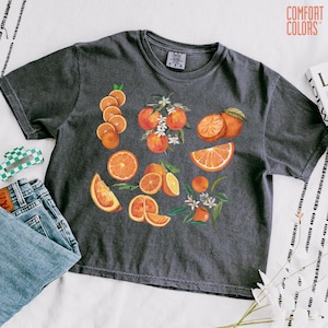 May include: Dark gray Comfort Colors t-shirt featuring a watercolor design of oranges, orange slices, and orange blossoms. The shirt is paired with blue jeans and a green and white checkered hair clip.