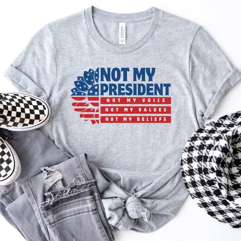 May include: Gray t-shirt with a red, white, and blue sunflower design. The text "Not My President" is printed on the shirt, along with the words "Not My Voice", "Not My Values", and "Not My Beliefs".