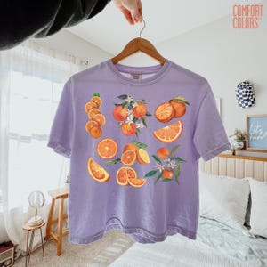 May include: A lilac Comfort Colors t-shirt featuring a vibrant orange fruit design. The graphic includes whole oranges, sliced segments, and blossoms. The shirt is hanging on a wooden hanger.