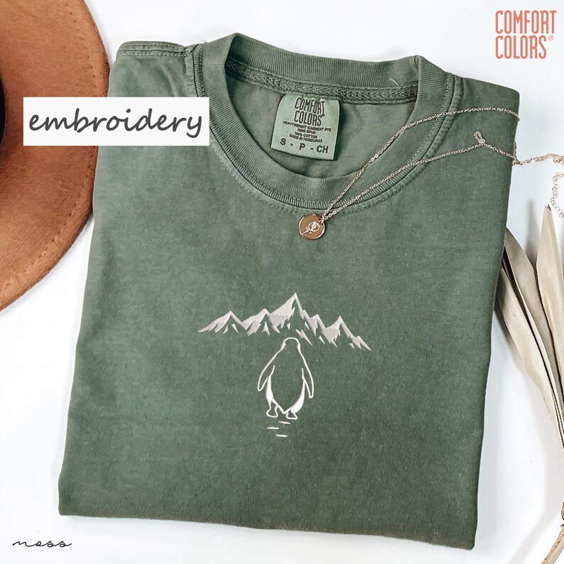 May include: An olive green t-shirt with an embroidered design of a penguin and mountains. The shirt has the word "embroidery" in a white box. A silver necklace with a pendant is on the shirt.
