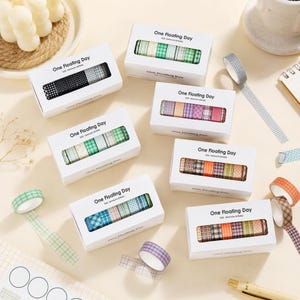 Puede incluir: Siete cajas rectangulares blancas etiquetadas como One Floating Day, cada una con seis rollos de cinta washi estampada. Las cintas presentan varios diseños de cuadros, dameros y lunares en verde, azul, morado, naranja y negro.