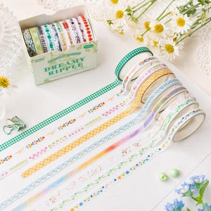 May include: A set of ten rolls of decorative washi tape in a cardboard box labeled Dreamy Ripple. The tapes feature various patterns including green gingham, floral motifs, yellow checks, and colorful stripes, displayed in a row on a white surface.