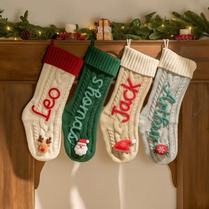 Peut inclure: Quatre bas de Noël en tricot torsadé, crème, vert, beige et gris, chacun avec un nom différent dans une couleur contrastante. Les bas sont décorés d'appliqués festifs comme un renne, un Père Noël et un bonbon. Une guirlande de lumières et de verdure est au-dessus.