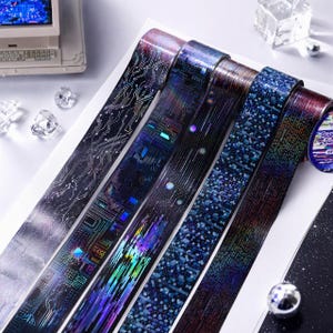Könnte beinhalten: Fünf Rollen dekoratives Klebeband mit holografischen und metallischen Designs. Die Muster zeigen komplexe Leiterplattenlinien, digitale Datenströme und pixelige Texturen in den Farben Dunkelblau, Violett und schimmerndem Silber.