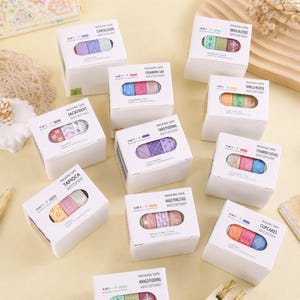 May include: A collection of ten white square boxes containing rolls of masking tape, each labeled with dessert-themed names like Cantaloupe, Strawberry Cake, Taro Pudding, and Cupcakes, featuring colorful patterned tapes visible through a cutout window.