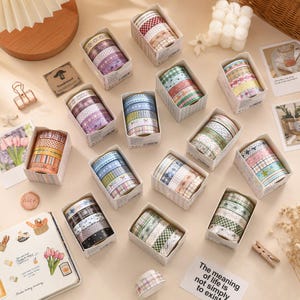 Op de afbeelding: Een verzameling decoratieve washi-tape rollen georganiseerd in kleine kartonnen doosjes, met diverse patronen waaronder ruiten, strepen, sterren en bloemmotieven in verschillende kleuren zoals roze, blauw, groen en paars, gerangschikt op een plat oppervlak.