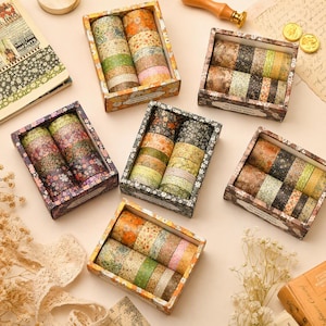 May include: Six rectangular boxes containing multiple rolls of decorative adhesive tape with various floral and botanical patterns in shades of green, orange, brown, and pink. Each box features a label with the text Blossoming on Deep Page.