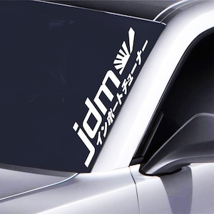 Può includere: Adesivo in vinile bianco con il testo "jdm" e il testo giapponese "インポートチューナー" (Import Tuner) sul parabrezza di un'auto nera e argentata.