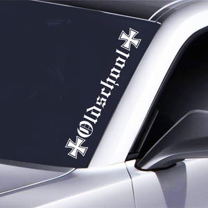 May include: White vinyl decal sticker with a black cross and the text "Oldschool" in a gothic font. The sticker is applied to the windscreen of a car.