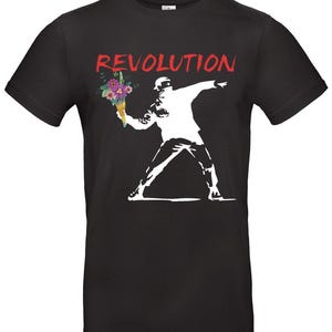 May include: Black t-shirt featuring a white graphic of a figure throwing a bouquet of flowers, with the word "REVOLUTION" in red above. The figure is wearing a mask and is in a throwing pose.