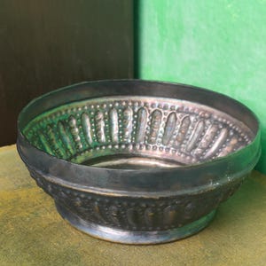 Puede incluir: Un cuenco decorativo de metal con un aspecto oscuro y envejecido. El cuenco presenta un patrón en relieve de formas verticales y puntos alrededor del borde y la base. El interior tiene un tinte verdoso y el cuenco se apoya en una superficie clara.