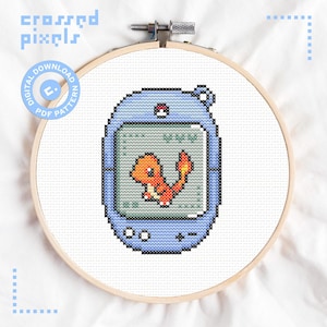 Puede incluir: Un patrón de punto de cruz en un bastidor de madera que muestra un dispositivo digital de mano de color azul claro con un personaje naranja pixelado de Charmander en la pantalla. El diseño incluye un pequeño botón circular y un mando de control.