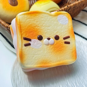 Könnte beinhalten: Eine Toastscheibe mit einem Katzengesicht-Design. Das Brot ist hellgelb mit orangefarbenen Akzenten, und das Katzengesicht hat braune Augen und Schnurrhaare. Der Toast liegt auf einem weißen Teller, mit anderen Backwaren im Hintergrund.