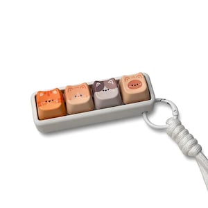 Peut inclure: Un ensemble de quatre touches de clavier sur le th&egrave;me des chats dans un plateau gris clair, chacune avec un design de visage de chat diff&eacute;rent. Les touches sont orange, beige, grise et p&ecirc;che. Une lani&egrave;re blanche est attach&eacute;e au plateau avec un fermoir argent&eacute;.