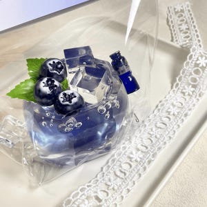 May include: A clear plastic bag containing a blue glass figurine of a blueberry drink with ice cubes and a small blue bottle. The figurine is sitting on a white surface with a white lace trim.