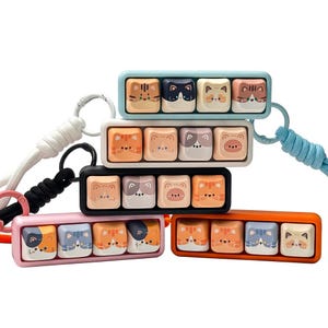 Peut inclure: Plusieurs ensembles de cinq touches de clavier, chacun avec un cadre de couleur diff&eacute;rente : bleu clair, blanc, noir, rose et orange. Chaque ensemble pr&eacute;sente cinq touches carr&eacute;es avec des motifs de visages de chats de diff&eacute;rentes couleurs et motifs. Les ensembles sont reli&eacute;s &agrave; des lani&egrave;res.