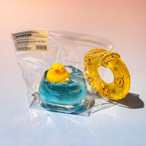 以下が含まれることがあります： 青いガラス容器に入った黄色いラバーダックのおもちゃ。透明なビニール袋に入っています。アヒルのイラストが入った黄色の浮き輪が袋の横にあります。袋にはテキストのラベルが付いています。