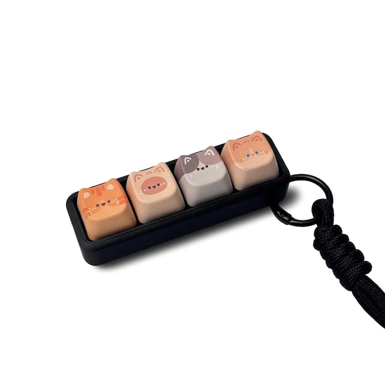 Peut inclure: Un ensemble de quatre touches de clavier sur le th&egrave;me des chats dans un plateau noir, chacune avec un design de visage de chat unique. Les touches sont orange, beige et grise. Une lani&egrave;re noire est attach&eacute;e au plateau.