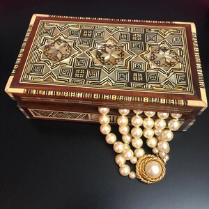 May include: A wooden box with intricate inlaid designs in shades of brown, gold, and white. The box is open and a pearl necklace with a gold clasp is resting on the edge.