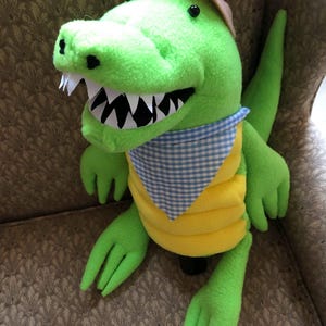 May include: A bright green plush alligator toy featuring a tan hat, a blue and white gingham neckerchief, and a yellow belly. The alligator has black button eyes and a wide open mouth with white felt teeth, showcasing a detailed, soft texture.
