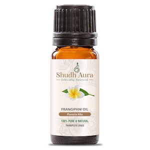 Op de afbeelding: Een klein amberkleurig glazen flesje met een zwarte dop, met het label "Shudh Aura Ethically Sourced" en "FRANGIPANI OIL Plumeria Alba 100% PURE & NATURAL THERAPEUTIC GRADE". Het label toont een witte frangipani-bloem met groene bladeren.