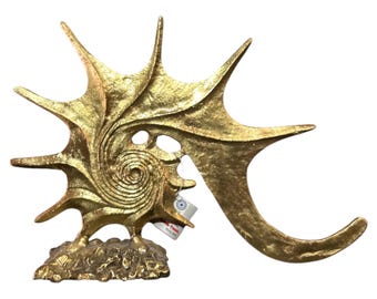 May include: A gold-colored metal sculpture of a seashell with a spiral design. The sculpture is on a textured base.