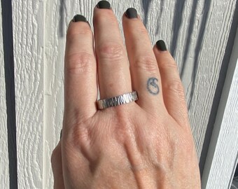 May include: A silver band ring with a textured hammered finish. The ring is worn on the middle finger of a hand.