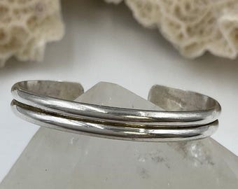 Pode incluir: Uma pulseira de punho prateada com um design de faixa dupla. A pulseira é exibida em uma superfície cristalina transparente, com um fundo texturizado esbranquiçado.
