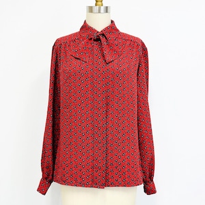 May include: A red long-sleeve blouse with a small black floral pattern. The blouse has a bow tie collar and is displayed on a mannequin. The blouse is a vintage style.