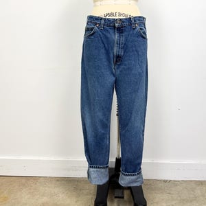 May include: Blue denim jeans with a high waist and a relaxed fit. The jeans have a classic five-pocket design, a button and zipper closure, and are cuffed at the ankles. The jeans are displayed on a mannequin against a white background.