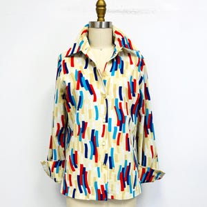 Puede incluir: Camisa abotonada con un estampado abstracto colorido. La camisa tiene un fondo crema con rayas verticales rojas, azules y beige. La camisa de manga larga se exhibe en un maniquí.