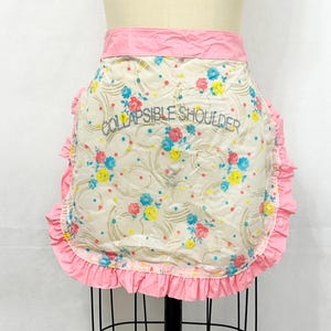 May include: A vintage, cream-colored apron with a pink ruffled trim and waistband. The apron features a floral pattern with blue, yellow, and pink flowers. The text "COLLAPSIBLE SHOULDER" is printed on the front.