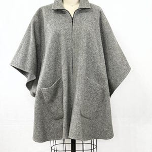 May include: A grey herringbone patterned cape featuring a high collar, a full-length front zipper closure, and two large patch pockets on the front. The garment has a loose, flowing silhouette with wide arm openings and a structured, tailored neckline.