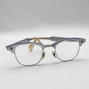 May include: A pair of vintage eyeglasses with a silver-colored frame and clear lenses. The glasses have a classic browline design with a subtle, angular shape. The temples are a light brown color.