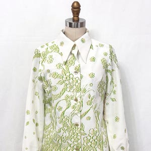 May include: White button-down shirt with a floral pattern in shades of green. The long-sleeved blouse features a wide collar and gold-colored buttons. The floral design covers the front and sleeves.