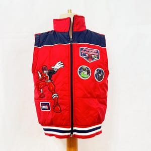 Puede incluir: Un chaleco acolchado rojo sin mangas con un panel superior azul oscuro y cremallera frontal. El chaleco presenta un gráfico grande de Power Rangers, parches bordados de personajes y el texto Power Rangers y Kicker en la parte delantera.