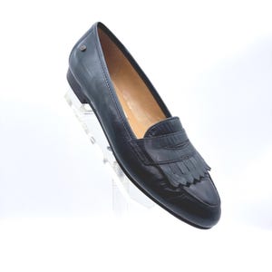 May include: A single black leather loafer with a decorative fringe detail. The shoe has a low heel and a rounded toe. The interior is a light tan color. The shoe is displayed on a clear stand against a white background.