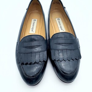 May include: A pair of black leather loafers with fringe detailing. The shoes have a rounded toe and a low heel. The brand name "Etienne Aigner" is visible on the insole. The shoes are displayed on a white background.