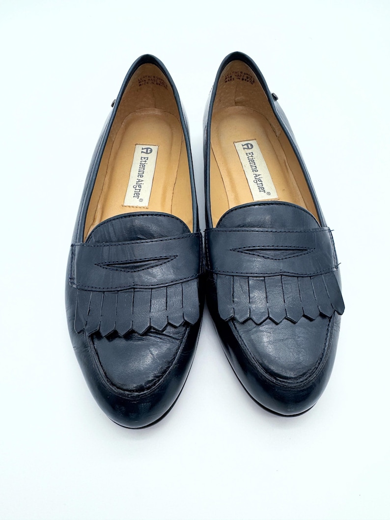 May include: A pair of black leather loafers with fringe detailing. The shoes have a rounded toe and a low heel. The brand name "Etienne Aigner" is visible on the insole. The shoes are displayed on a white background.
