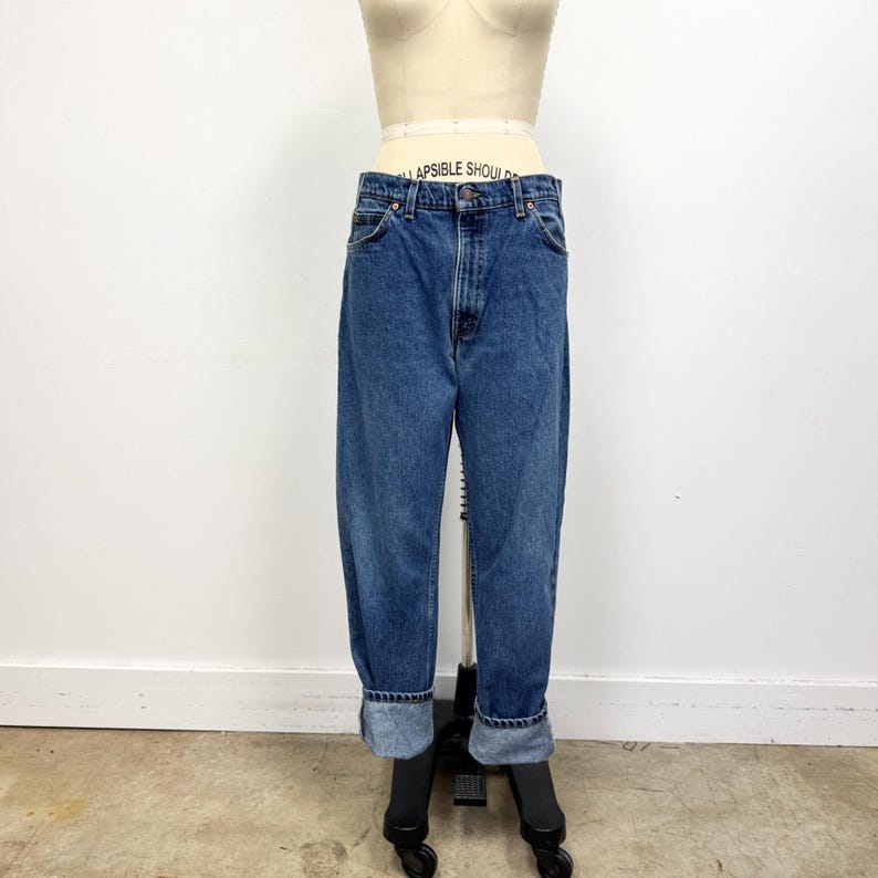 May include: Blue denim jeans with a high waist and a relaxed fit. The jeans have a classic five-pocket design, a button and zipper closure, and are cuffed at the ankles. The jeans are displayed on a mannequin against a white background.