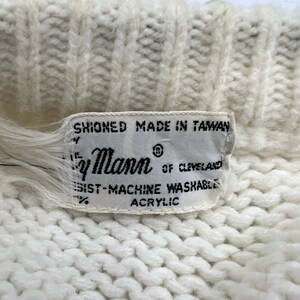K&ouml;nnte beinhalten: Wei&szlig;er Strickpullover mit einem wei&szlig;en Etikett mit der Aufschrift "Shioned Made in Taiwan" und "By Mann of Cleveland" mit dem Text "100% Machine Washable Acrylic".