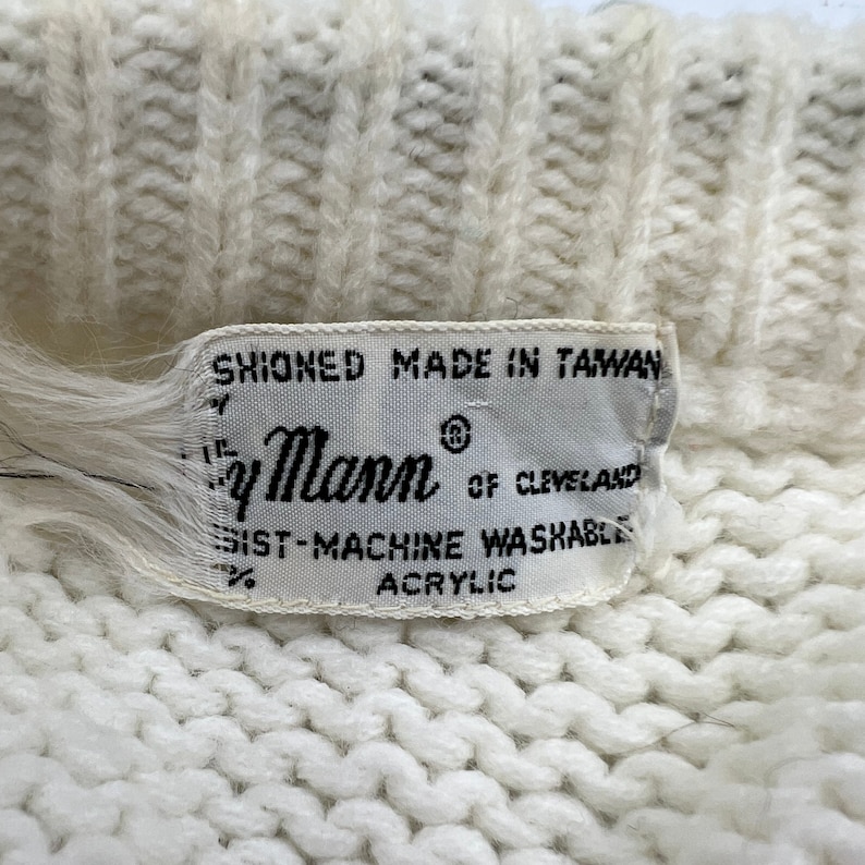K&ouml;nnte beinhalten: Wei&szlig;er Strickpullover mit einem wei&szlig;en Etikett mit der Aufschrift "Shioned Made in Taiwan" und "By Mann of Cleveland" mit dem Text "100% Machine Washable Acrylic".