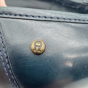 May include: Close-up of a navy blue leather loafer. The shoe features a gold-colored emblem and blue stitching details. The leather has a textured appearance, and the stitching is visible along the seams.